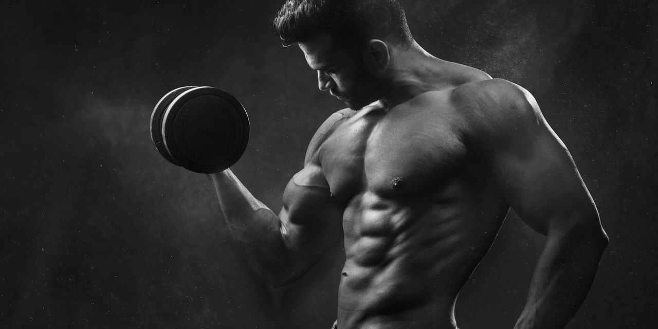 Powerful black and white portrait of a shirtless bodybuilder lifting dumbbells in the gym.