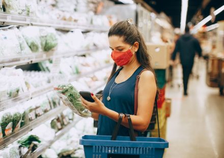 A woman wearing a mask shops for groceries in a supermarket, checking products with her phone.