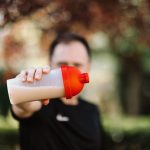 Focus on a man holding a plastic protein shake tumbler outdoors with a blurred background.