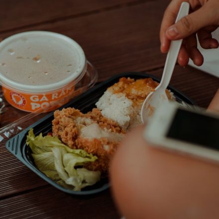 A hand holding a spoon near a meal of fried chicken, rice, and soup.